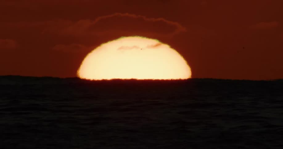 A close-up shot of the sun's distorted golden orb sinking below the ocean horizon under a deep red sky
