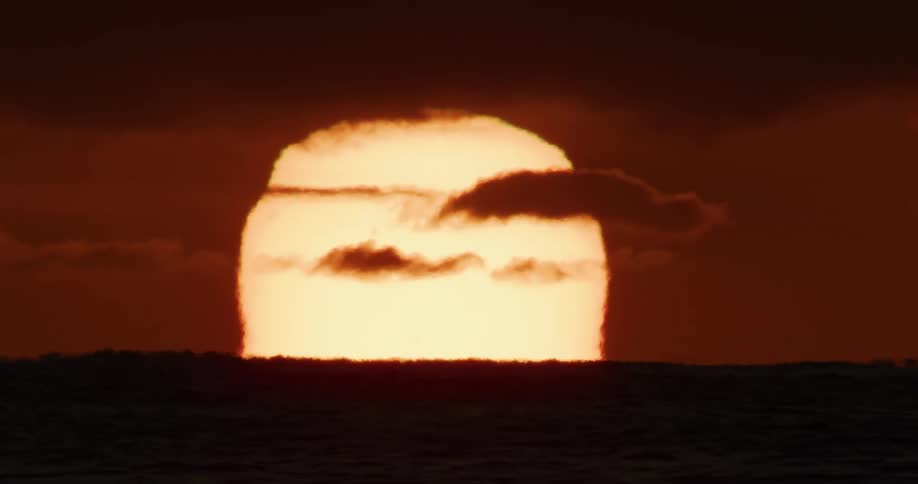 A powerful telephoto shot shows the huge, glowing sun sitting on the horizon as dark clouds pass over the ocean