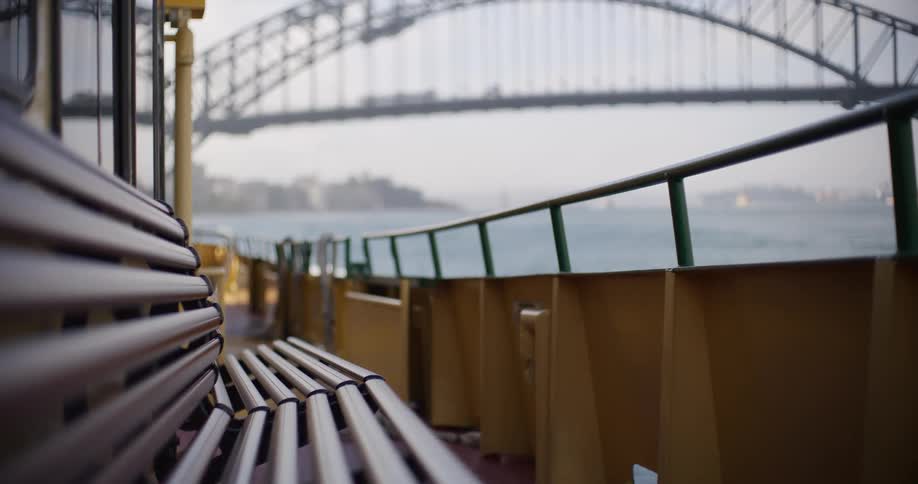 An empty bench on a ferry provides a serene view of the Sydney Harbour Bridge on a hazy day