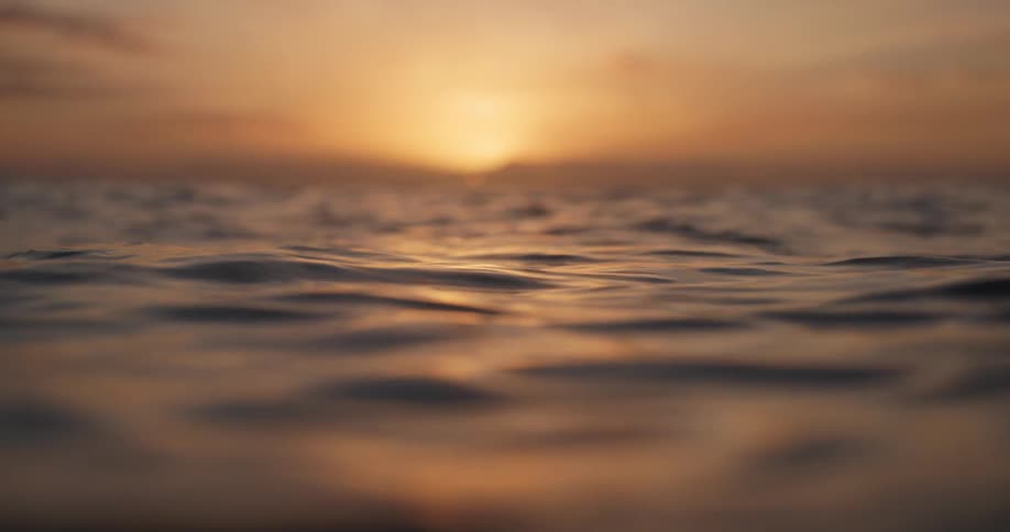 A close-up, low-angle slow motion shot of calm ocean waves at sunrise with a beautiful golden glow on the horizon