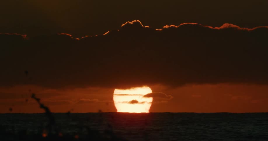 A close-up of the bright sun setting over the ocean, peeking from underneath a thick layer of dark clouds
