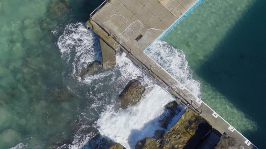 Dramatic slow-motion top-down shot of powerful ocean waves crashing over a rock pool barrier on a sunny day