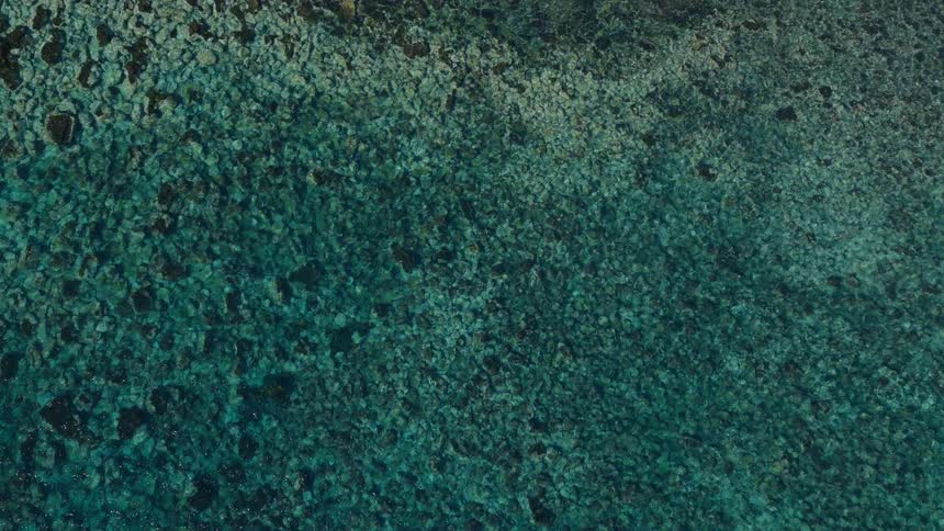 A top-down aerial view of calm, clear turquoise ocean water revealing a rocky seabed below