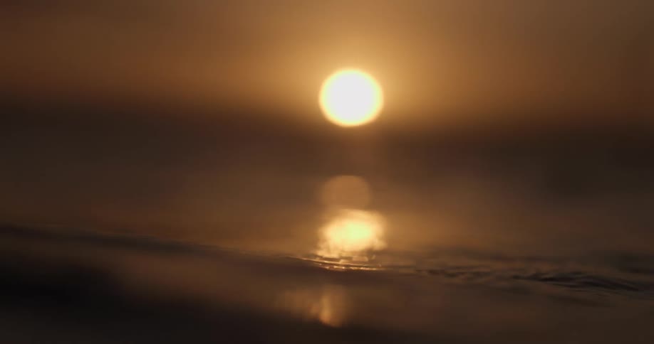 Calm, misty water ripples in slow motion as the golden sun sets on the horizon, its reflection shimmering on the surface