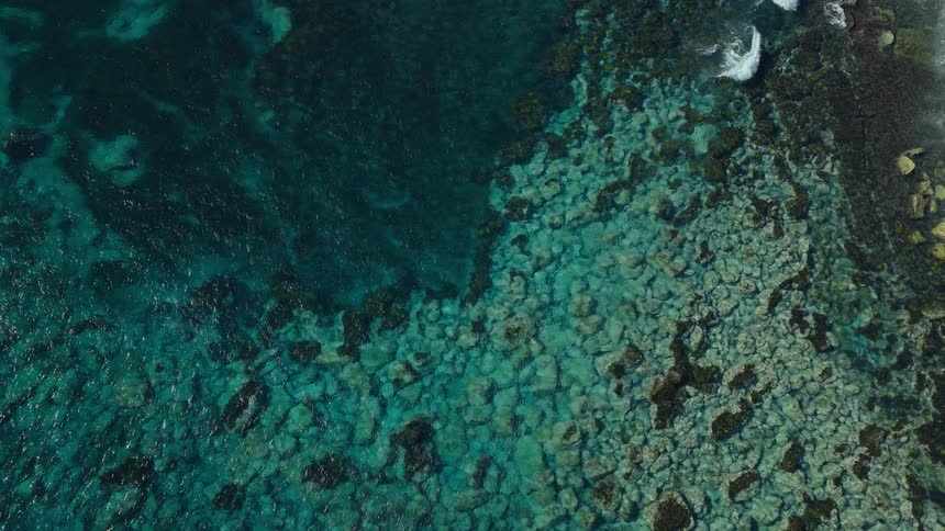 A serene top-down aerial view of the clear turquoise ocean lapping against a shallow rocky reef