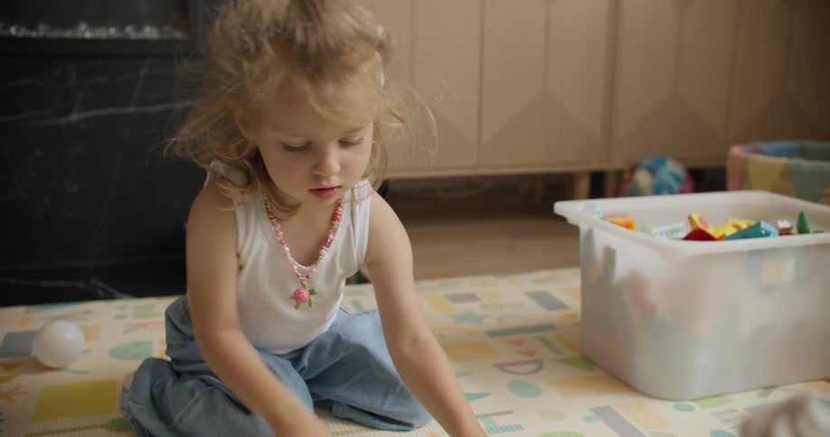 A cute toddler with blonde hair sits on a playmat in her living room, focused on her toys