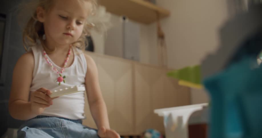 A cute little girl with a beaded necklace concentrates intently while playing with building blocks in her room
