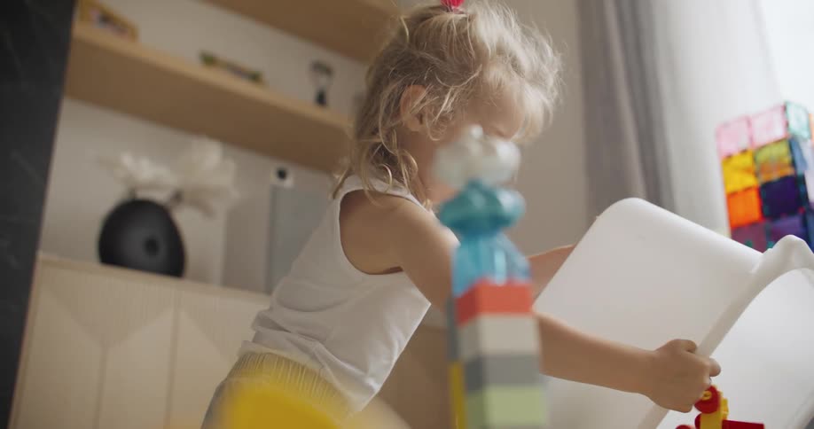 A cute toddler with messy blonde hair is focused on emptying a bin full of toys in her playroom in slow motion