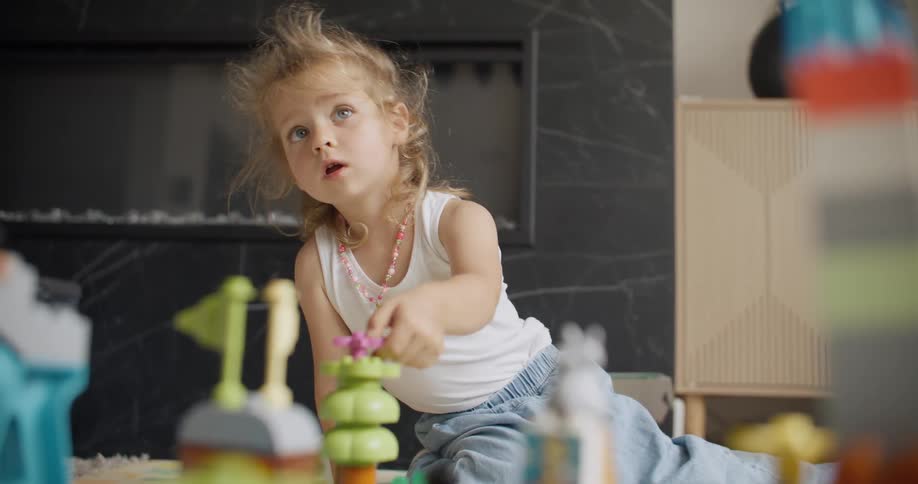 An adorable little girl with blonde hair sits on the floor and plays with colorful plastic building blocks in a modern living room
