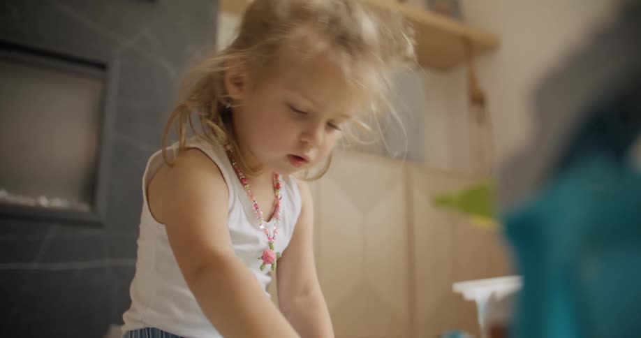 A cute little blonde girl with a pink necklace sits on the floor, deeply focused on playing with her toys