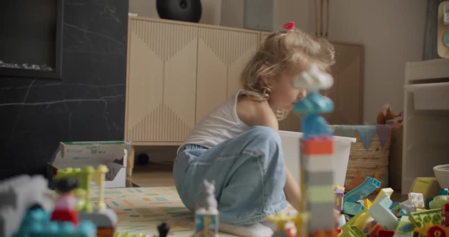 An adorable little girl sits on the floor of her playroom, focused on building with colorful construction blocks