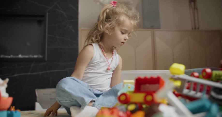 A cute little blonde girl sits on the living room floor, focused on playing with her many colorful toys