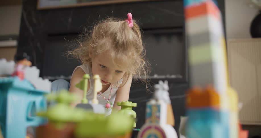 A cute little blonde girl with a pink hair tie concentrates deeply while playing with her colorful plastic building blocks