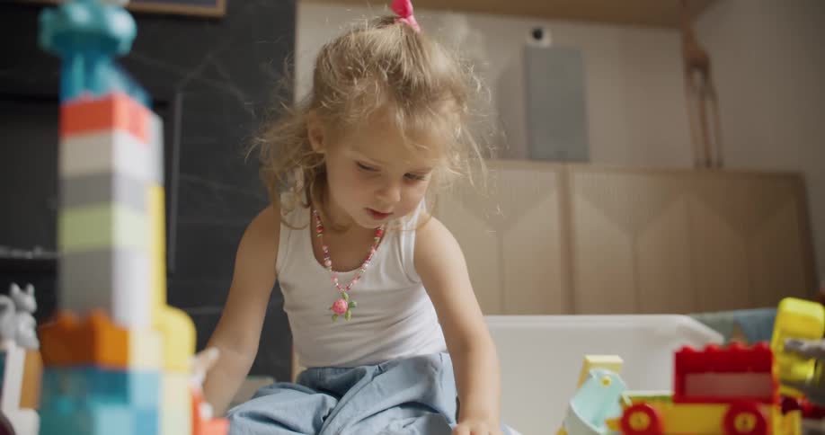 A cute little girl with curly blonde hair plays with colorful building blocks in her room