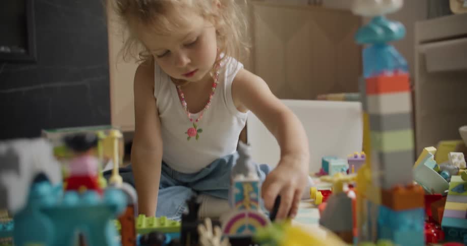 A cute little blonde girl sits on the floor, deeply engrossed in building structures with her colorful toy blocks in slow motion