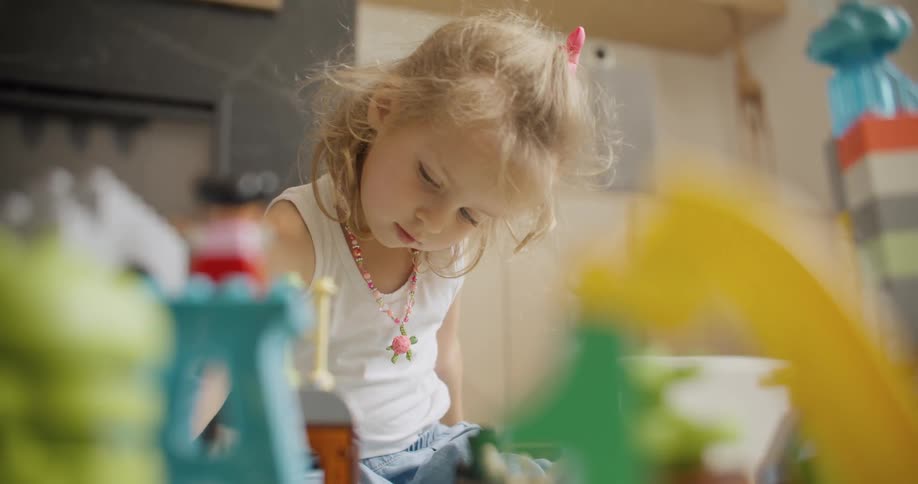A cute little blonde girl is deeply focused on playing with her colorful toys and building blocks in her room