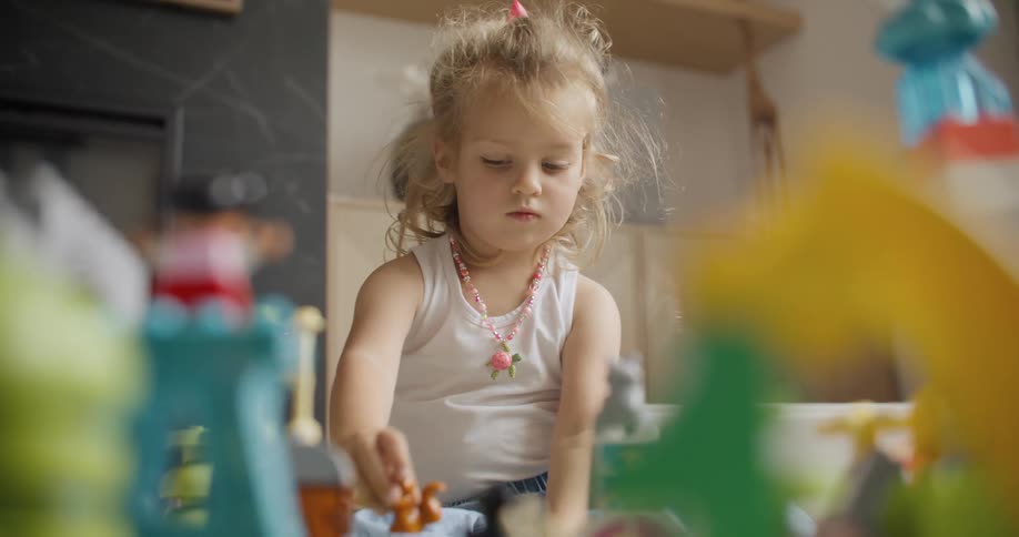 A cute little blonde girl with a pink necklace is engrossed in playing with her colorful building blocks and figurines on the floor