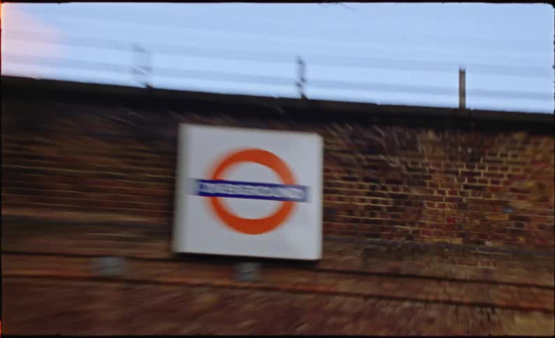 A wobbly, handheld shot captures the iconic London Overground sign on a brick wall, filmed with a vintage Super 8 aesthetic