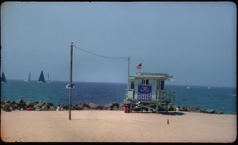 Vintage film captures a sunny day at a California beach with a lifeguard tower and sailboats on the water