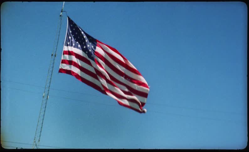 A vintage handheld shot from a moving vehicle captures American flags waving against a clear blue sky