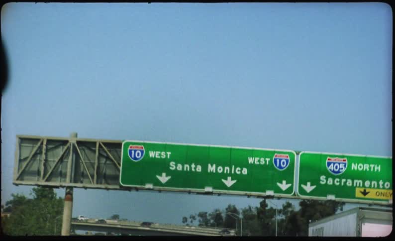 A vintage, handheld point-of-view shot from a car driving on a Los Angeles freeway towards a Santa Monica sign