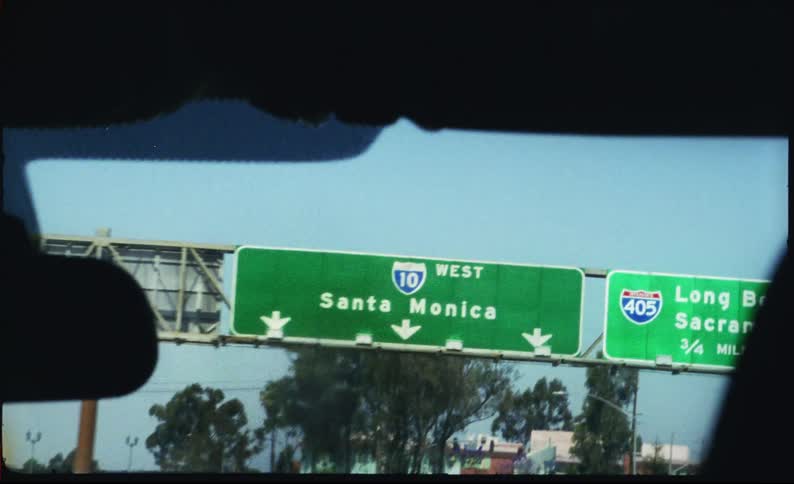 A vintage, handheld shot from a car driving on a Los Angeles freeway towards a Santa Monica highway sign