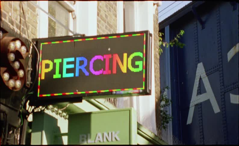 A colorful electronic sign outside a tattoo and piercing parlor flashes its services in rainbow pixelated lettering