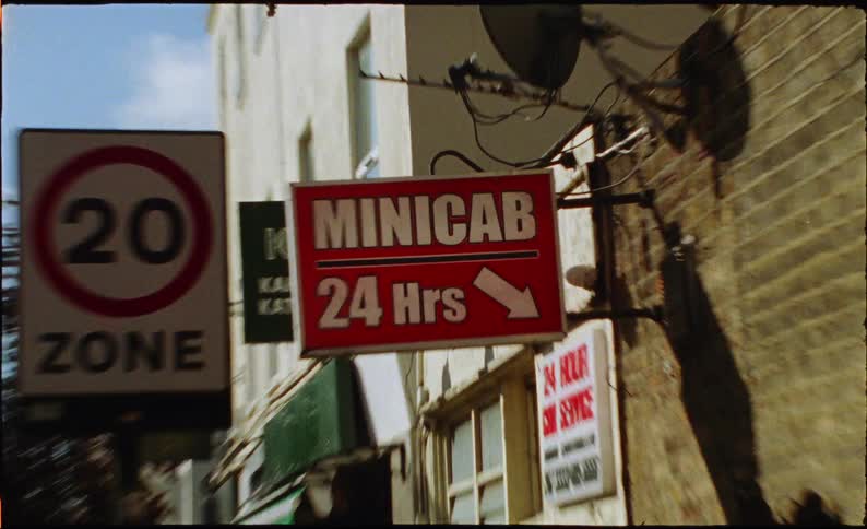 A grainy, film-style shot zooms in on a bright red 'Minicab 24 Hrs' sign next to a speed limit sign on a sunny London street