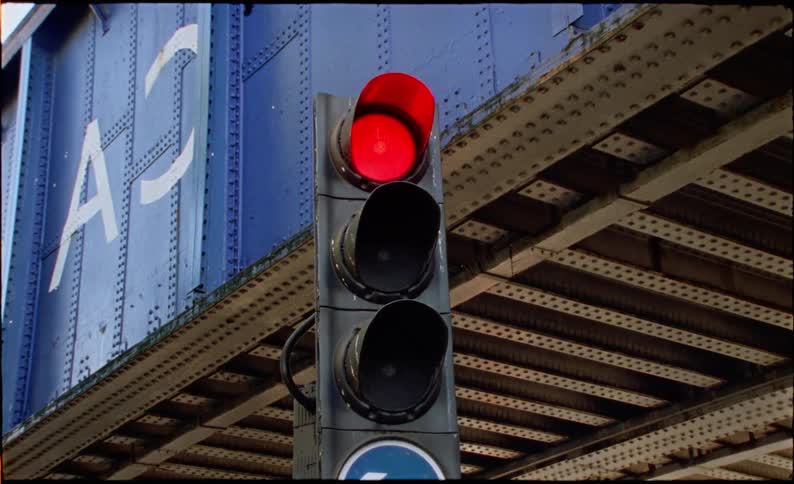 A traffic light cycles from a green arrow to red in a grainy, low-angle shot beneath an industrial city bridge