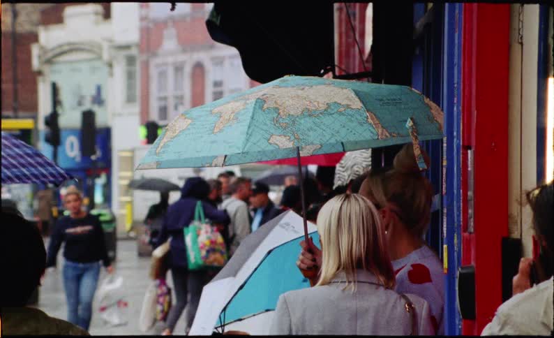 In a slow-motion, film-style shot, pedestrians navigate a rainy city street with umbrellas, with a close-up on a unique world map design