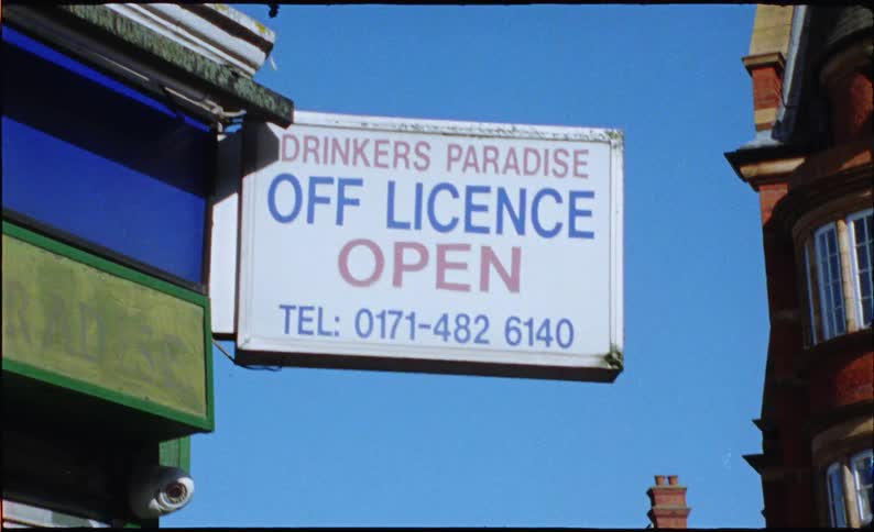 A vintage film shot captures a low-angle view of a 'Drinkers Paradise' off-licence sign on a sunny London street corner