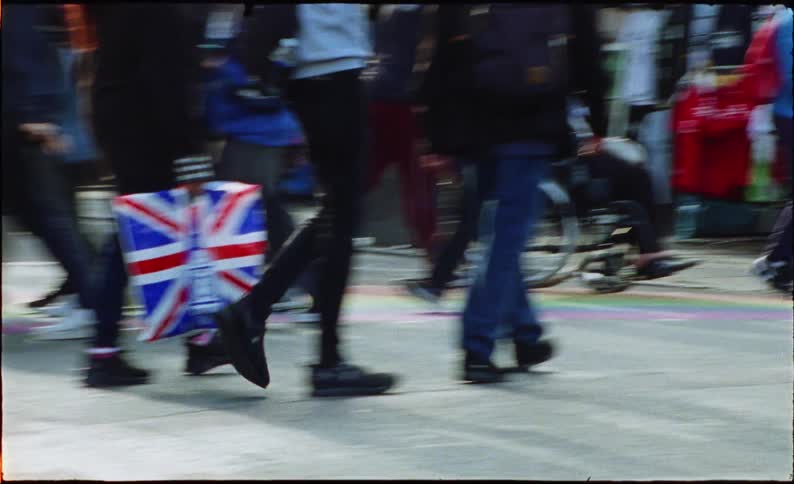 A low-angle, vintage-style shot captures the bustling feet of a diverse crowd walking over a rainbow crosswalk on a busy city street