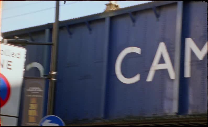 A vintage film-style shot of the iconic Camden Town bridge sign, with a red double-decker bus and city traffic passing by on a bustling London street