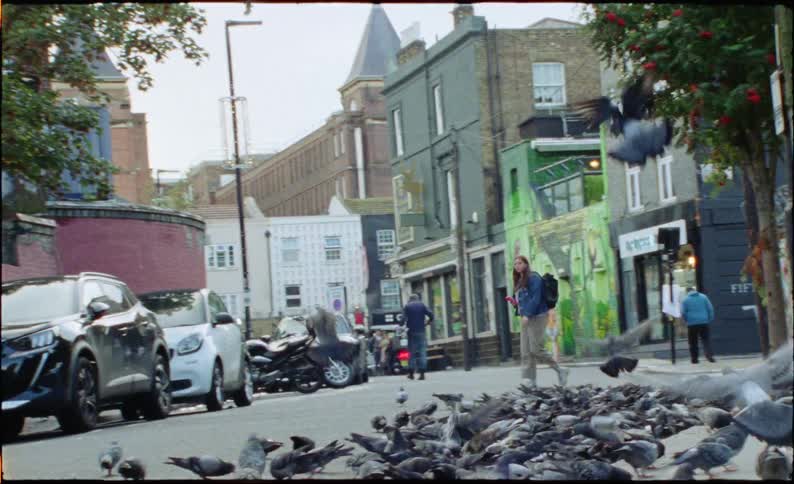A blurry, slow-motion handheld shot of a man on a scooter riding through a large flock of pigeons on a city street, causing them to fly up in a chaotic flurry