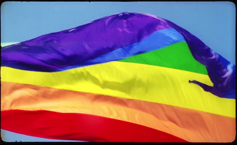 A vibrant rainbow Pride Flag waves in slow motion against a clear blue sky, captured on vintage-style film