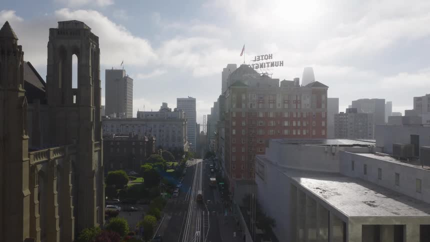 A slow aerial reveal shows a San Francisco cable car on a downtown street, with the city skyline rising in the hazy background