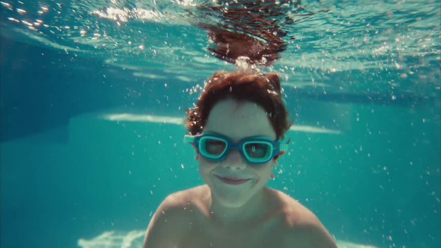 Young boy wearing blue goggles smiles underwater in a sunny swimming pool