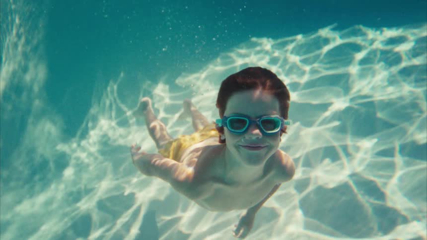 A happy young boy with red hair and goggles smiles as he swims towards the camera underwater in a sunlit pool in slow motion