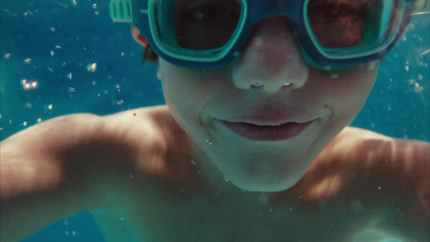 A happy young boy wearing goggles swims underwater, smiling directly at the camera in a sunlit pool