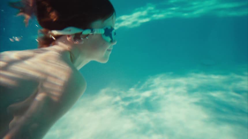 A smiling boy with red hair and goggles enjoys a slow-motion swim underwater in a beautiful sunlit swimming pool