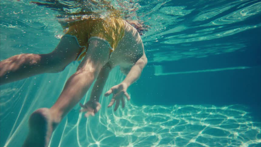 Slow motion underwater view of a young boy in yellow swim trunks swimming gracefully and doing a flip in a sparkling pool
