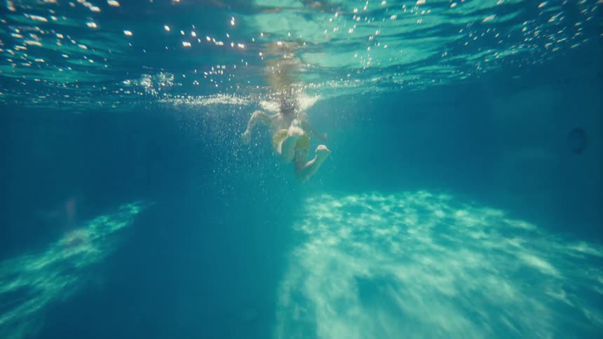 A young boy in yellow shorts floats and kicks his legs in slow motion, seen from underwater in a sunlit swimming pool
