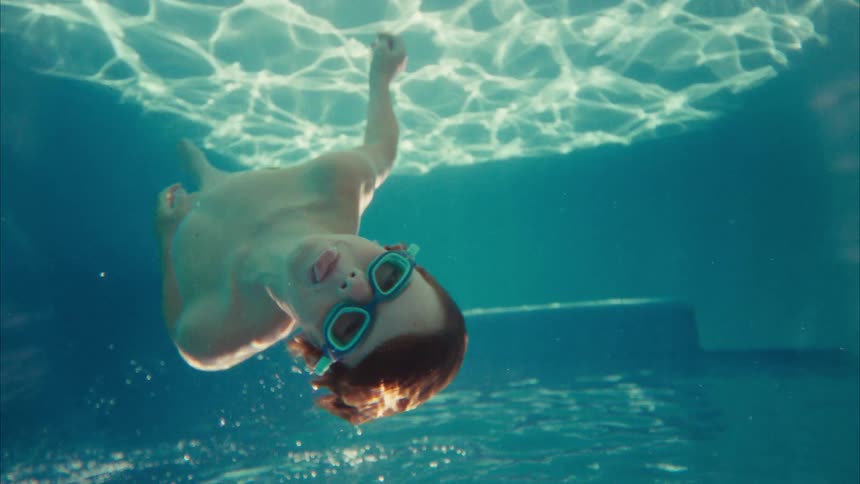A happy young boy wearing goggles floats upside down while swimming underwater in a pool