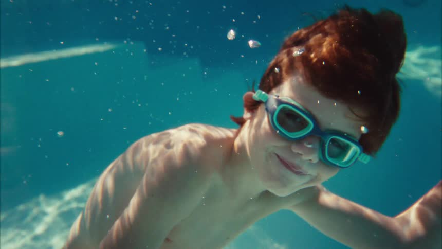 A happy red-haired boy wearing goggles swims gracefully underwater in a sunlit pool, smiling for the camera
