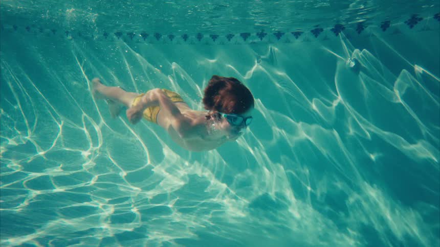 A young boy with goggles and yellow swim trunks swims gracefully underwater in a bright blue swimming pool