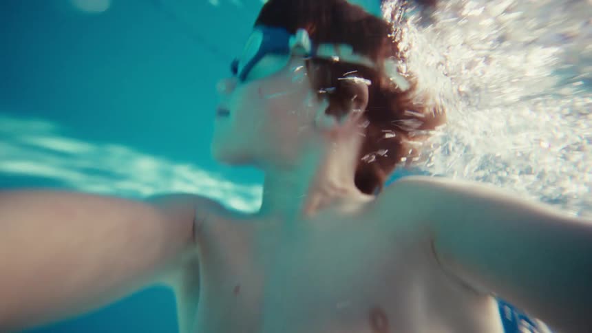 A joyful young boy wearing swim goggles films himself swimming underwater in a bright blue swimming pool