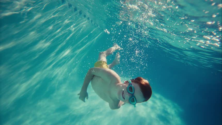 A slow-motion underwater view captures a happy red-haired boy with goggles gracefully swimming towards the camera in a sunlit pool