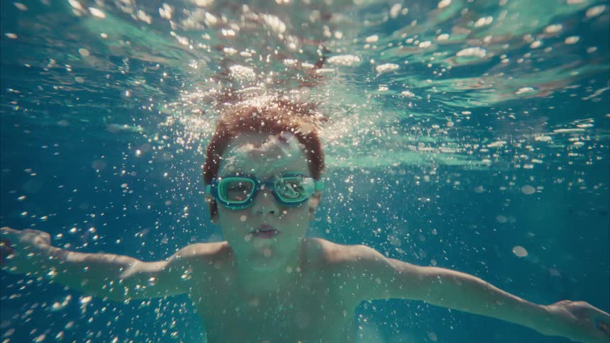 A young boy with goggles floats peacefully underwater in a sunlit swimming pool, looking at the camera in slow motion