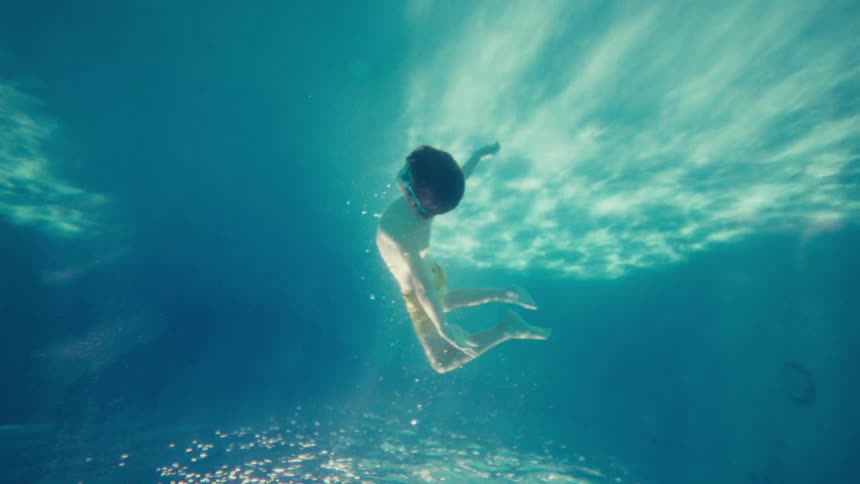 From an underwater perspective, a young boy with goggles glides through the shimmering blue water in slow motion