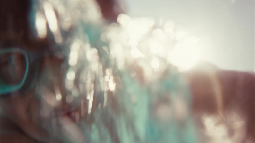 A smiling young boy in swim goggles plays in the water, captured in a slow-motion, point-of-view shot as he goes underwater and resurfaces